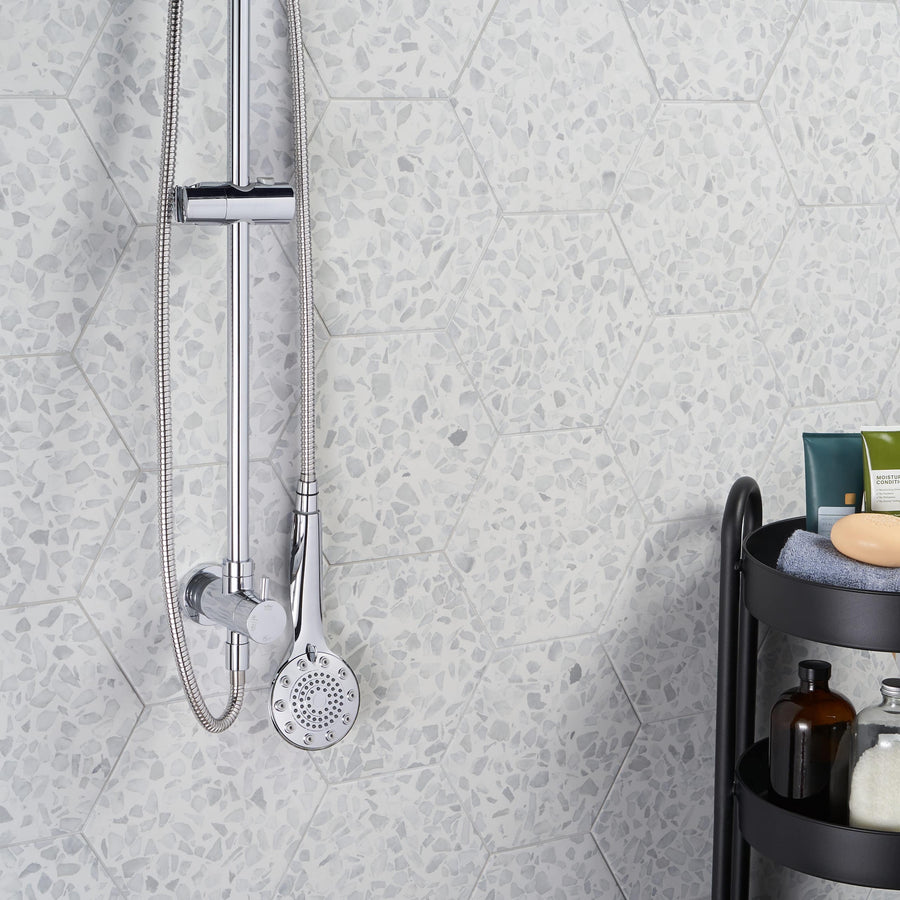 A modern shower with a chrome-finished detachable showerhead is mounted on a wall of Riazza 9" Hexagon Porcelain Tile. Nearby, a black shelf holds assorted bottles, soap, and a folded towel.