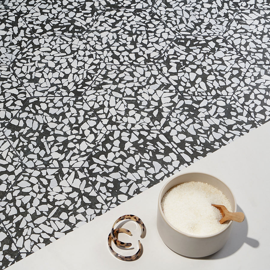 A bowl of white bath salts with a wooden scoop sits beside a marbled hair clip, set against a chic black and white Riazza 9" Hexagon Porcelain Tile background for a modern touch.