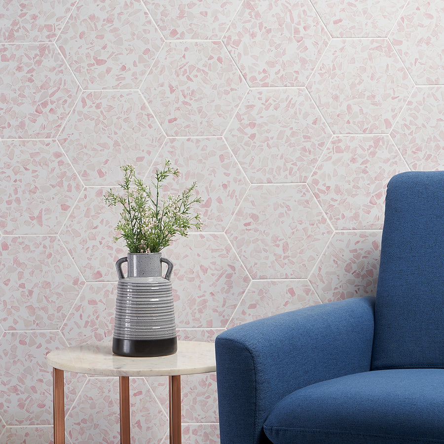 A small marble table with a gray striped vase and green branches stands by a blue upholstered armchair. The background features Riazza 9" Hexagon Porcelain Tile in pink and white, adding charm with contemporary floor and wall tiles.