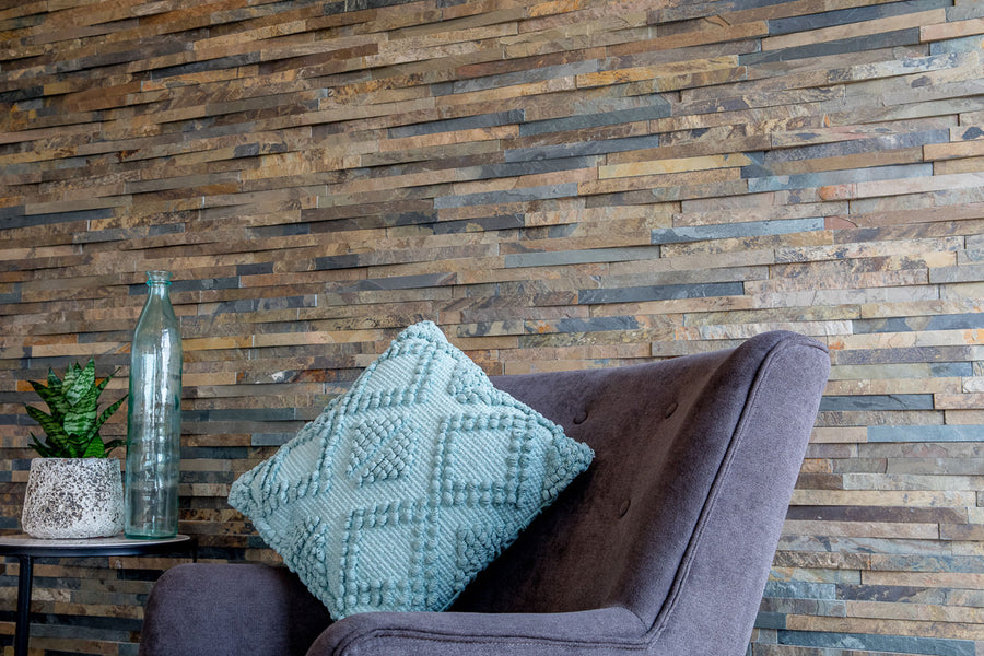 A modern gray armchair with a textured blue cushion sits beside a table with a plant and glass bottle, set against a Rustic Cladding 4x22 3/4 Natural Stone Ledger Panel backdrop that enhances the cozy, contemporary vibe.