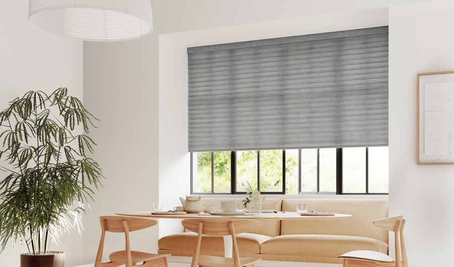A modern dining nook features a round wooden table, four chairs, a beige cushioned bench, a large potted plant, and a window fitted with Sonnette™ Cellular Roller Shades that filter natural light.
