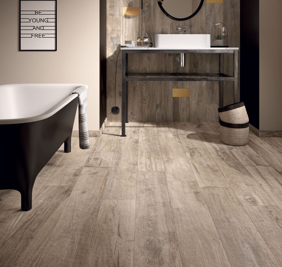 A modern bathroom featuring Legend 8x48 Wood Plank Tile flooring, a black and white freestanding tub, round mirror over the sink, striped towel, woven baskets, and a "BE YOUNG AND FREE" sign creates a fresh wood-inspired look.