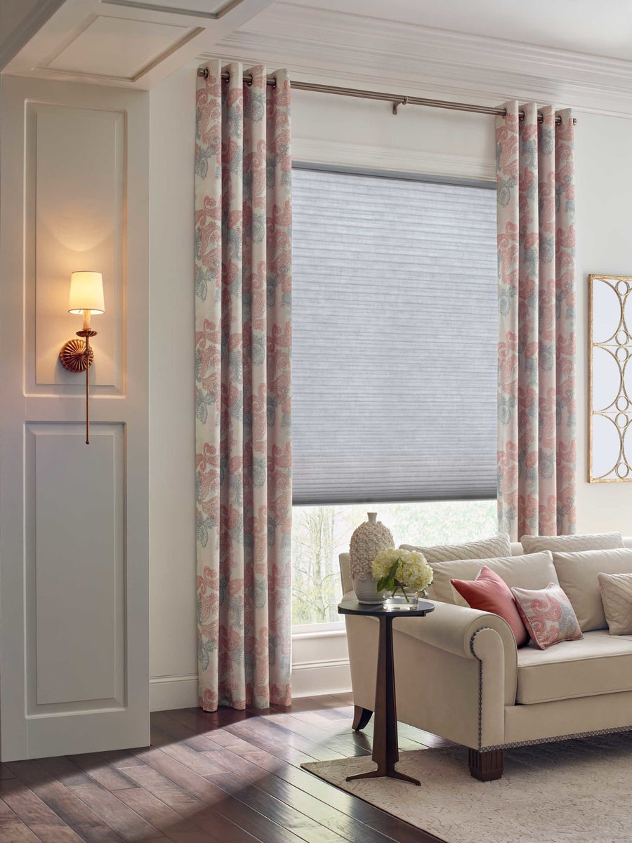 A bright living room with floral curtains, Duette® Honeycomb Shades for energy-efficient windows, a white sofa with pink and beige pillows, a small side table topped with a vase of flowers, and a lit wall sconce on a paneled wall.