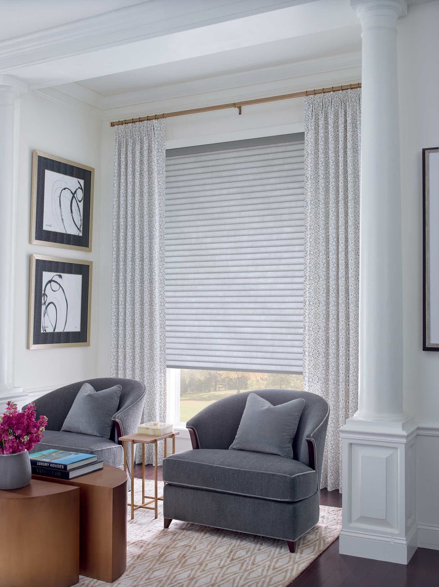 A bright, elegant living room featuring two gray armchairs, light-patterned curtains, a large window with Sonnette™ Cellular Roller Shades, abstract wall art, a side table, and a coffee table topped with a vase of pink flowers.