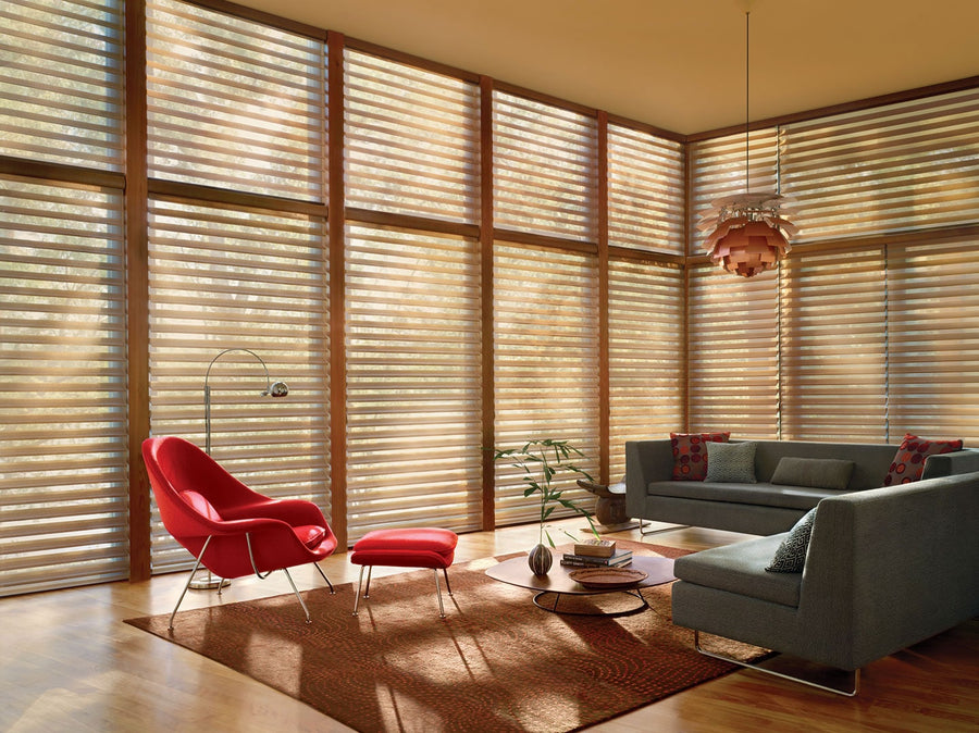 A modern living room with large windows covered by Smart Blinds & Shades. The space has a red lounge chair with ottoman, two gray sofas, a round coffee table, floor lamp, and a patterned brown rug.