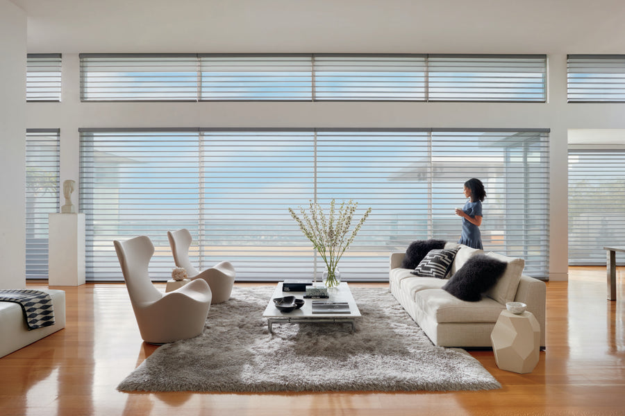 A modern living room with large windows, neutral furniture, a gray rug, and decorative accents. Silhouette® Window Shadings with PowerView® Motorization softly diffuse light as a woman stands by the window holding a cup.
