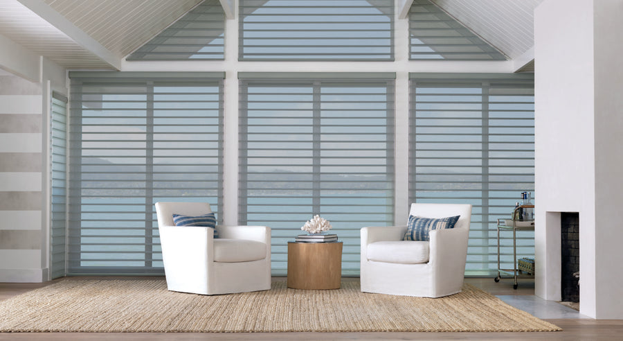 A bright, modern living room with two white armchairs, a round wooden table, a woven rug, and large windows featuring Silhouette® Window Shadings with PowerView® Motorization for gentle light control and scenic water views.