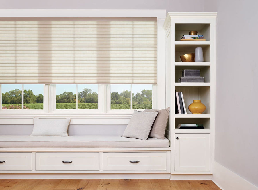 A built-in window seat with light cushions and pillows sits below three large windows fitted with Sonnette™ Cellular Roller Shades. Next to it is a white bookshelf holding books, decor, and a yellow vase. The floor is light wood.