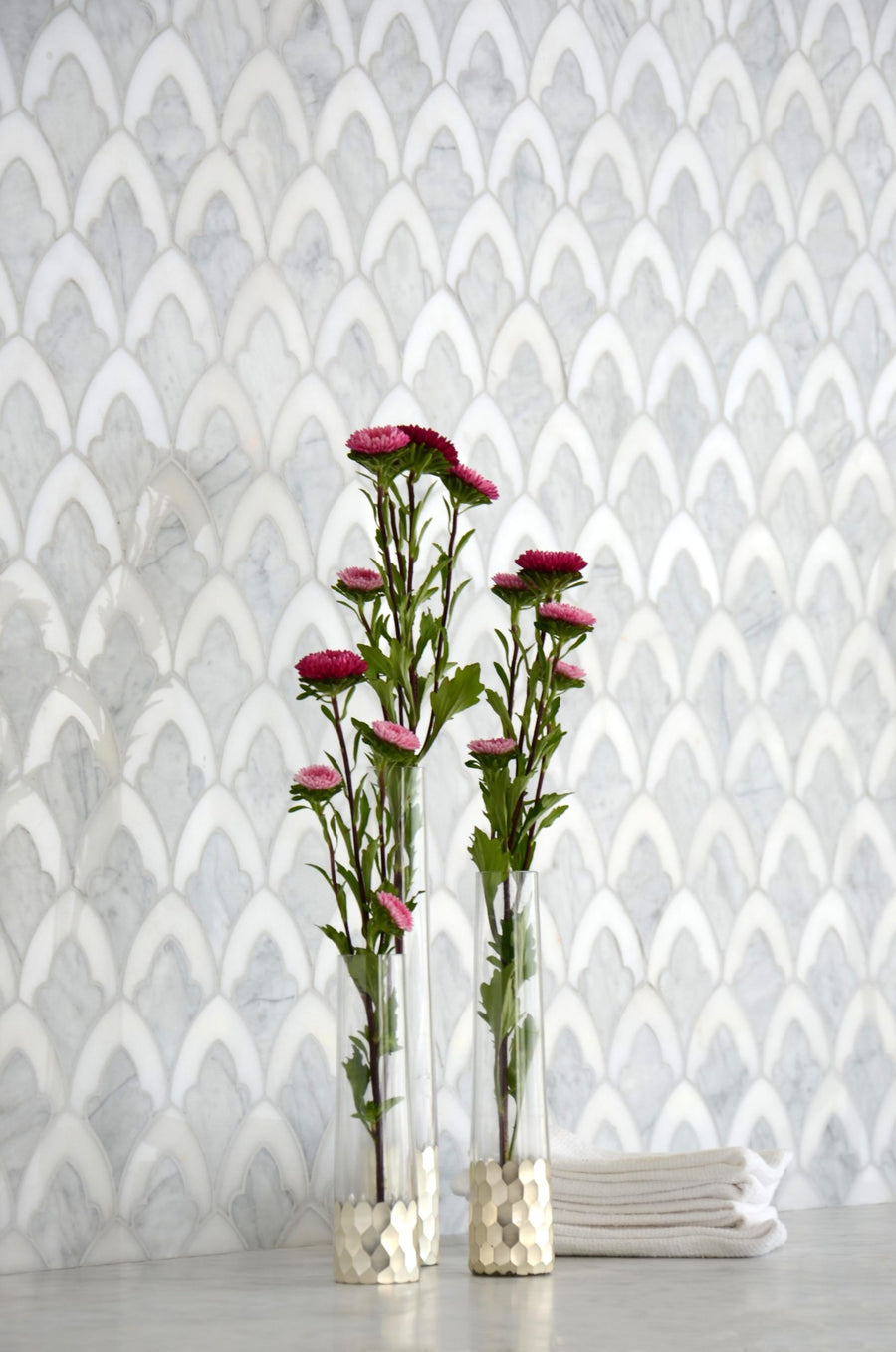 Two clear vases with gold bases holding pink flowers sit on a light surface in front of a decorative wall featuring Talya Sophia 8 3/4 x 13 1/2 Waterjet Natural Stone in a fish scale pattern, with folded white towels stacked in the background.