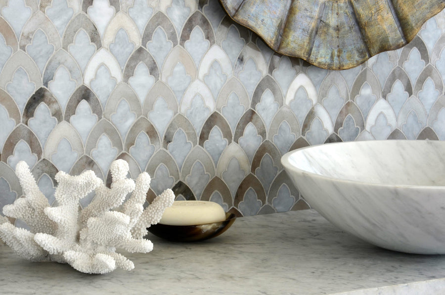 A close-up of a bathroom countertop featuring a white coral decoration, round soap dish, marble bowl sink, and Talya Sophia 8 3/4 x 13 1/2 Waterjet Natural Stone tiles as a decorative backsplash in neutral tones.