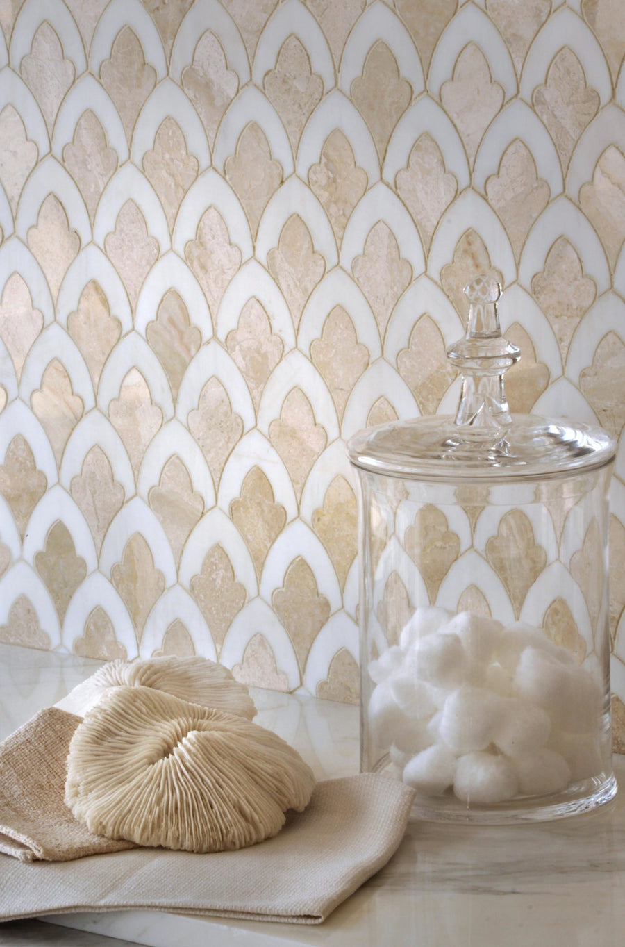A glass jar with cotton balls sits beside beige towels and a natural sponge on a marble surface, set against modern cream and white walls featuring Talya Sophia 8 3/4 x 13 1/2 Waterjet Natural Stone tiles.