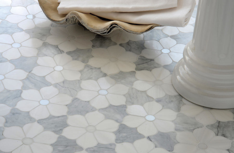 Close-up of a Talya Theodora 12 1/8 x 14 Waterjet Natural Stone tile floor with a white floral pattern on a light gray background. A folded cloth sits in a shell-shaped dish next to a cylindrical white ceramic piece.