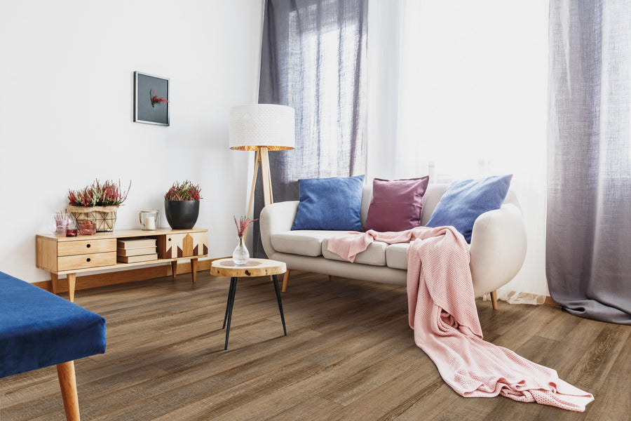 Bright, modern living room featuring a white sofa, colorful pillows, pink throw, waterproof COREtec Everyday flooring, side table, floor lamp, plants, mid-century console, and sheer curtains letting in natural light.