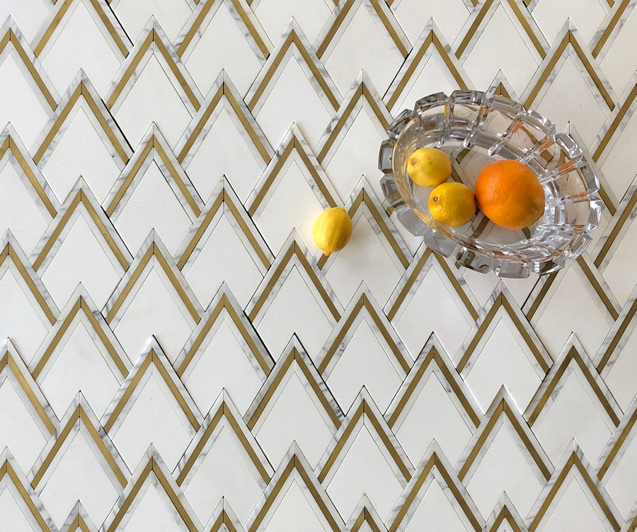 A glass dish holding an orange and two lemons sits on a white, zigzag-patterned surface inspired by Avenza Diamond Peak Calacatta Gold with Brass Marble; another lemon rests outside the dish on this elegant pattern.