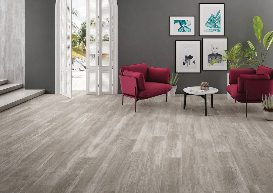 A modern living room features Driftwood II 8x48 Wood Plank Tile flooring, gray walls, red armchairs, a round coffee table, green plants, and framed art. Large arched windows offer sunny views of palm trees outside.