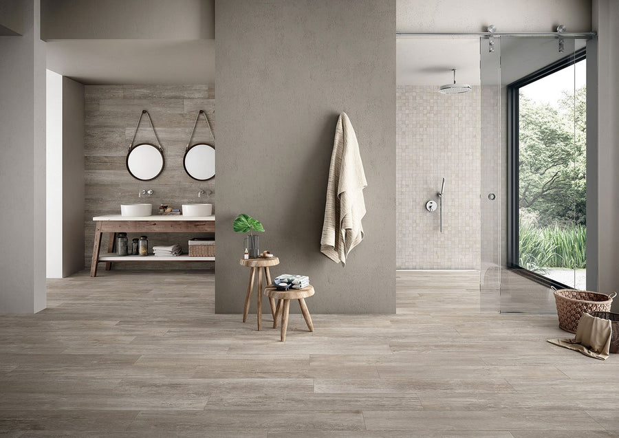 Modern bathroom with neutral tones featuring double sinks with round mirrors, a walk-in shower with glass doors and textured tile, Driftwood II 8x48 Wood Plank Tile, wooden stools, towels, large window, and green outdoor view.