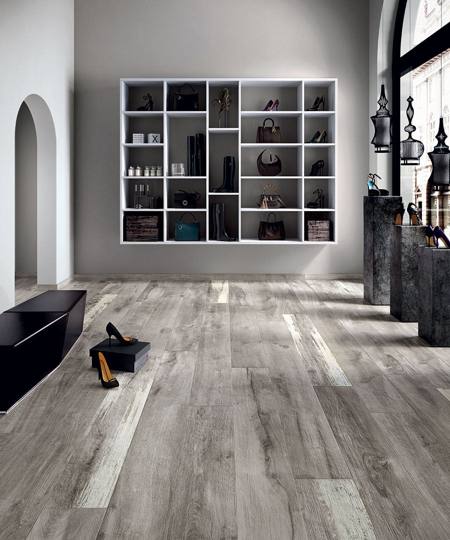 A modern shoe store featuring Legend 8x48 Wood Plank Tile in gray tones, white shelves for shoes and bags, large arched windows bringing in natural light, and shiny black benches with a few shoes on the floor.