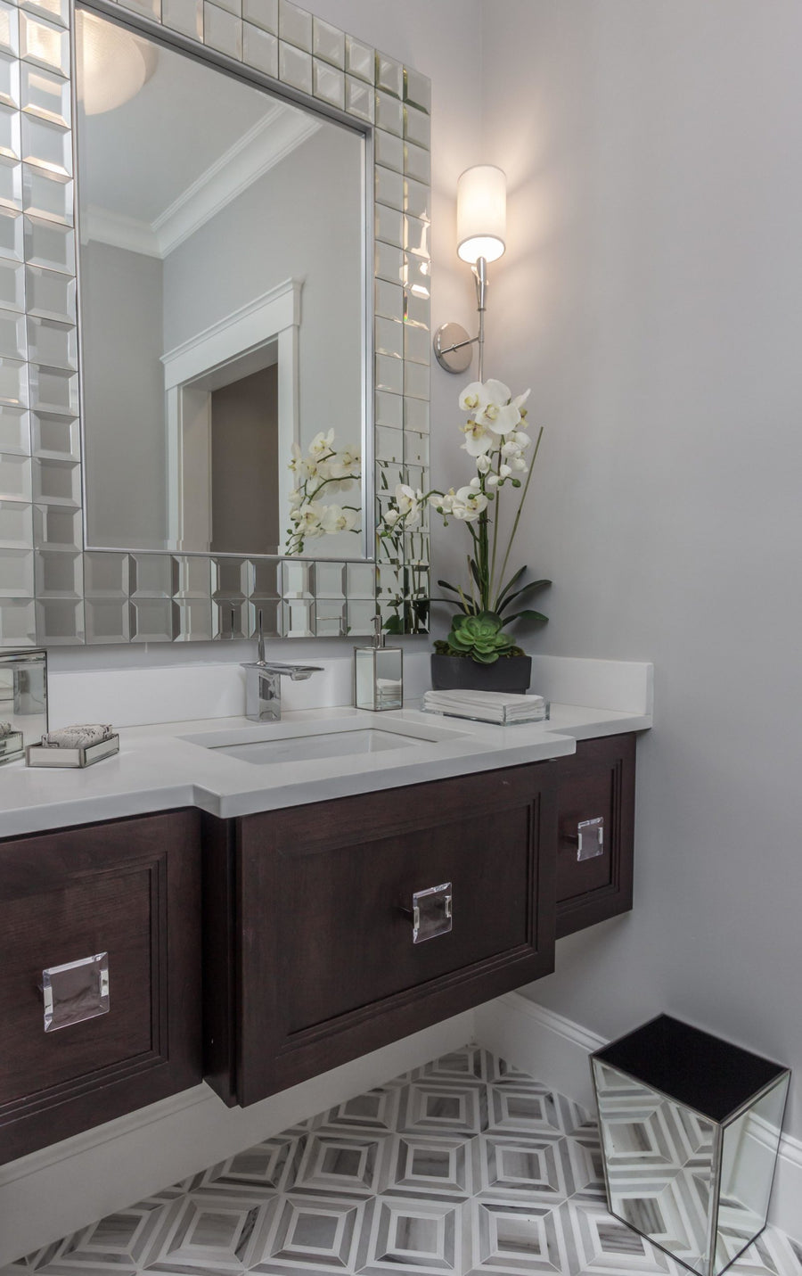A modern bathroom features Talya Yildiz 8 13/16 x 11 Waterjet Natural Stone geometric floor tiles, a dark wood vanity, and a large mirror. Marble and stone accents, plus an orchid in a rectangular vase, add an elegant touch.