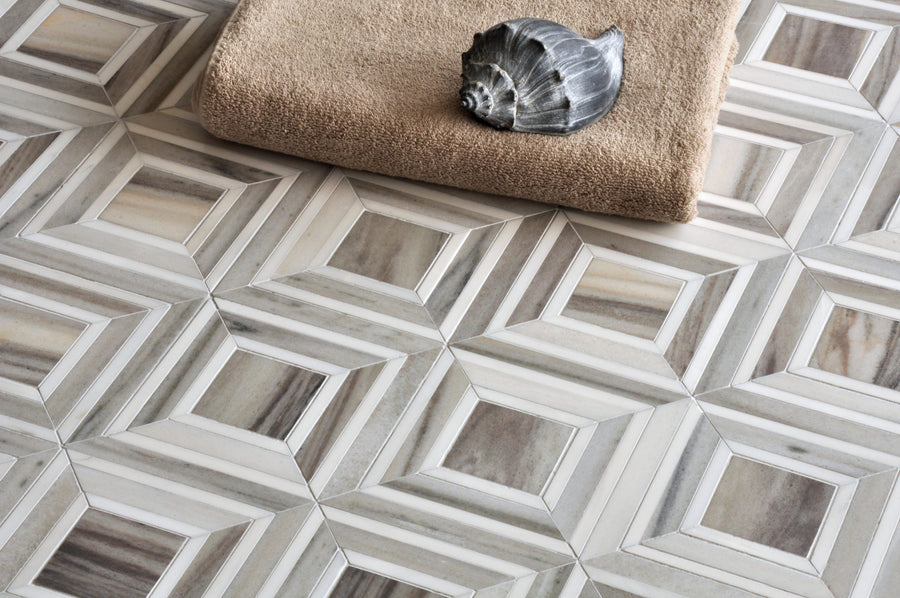 The Talya Yildiz 8 13/16 x 11 Waterjet Natural Stone tile showcases modern gray and white geometric patterns, resembling marble and stone. A folded beige towel and a metallic seashell ornament rest on the surface.