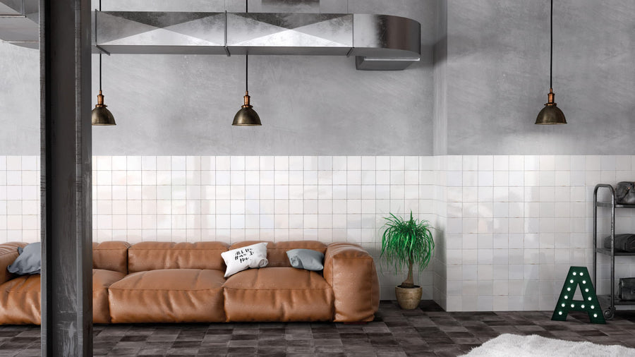 A modern living room with a brown leather sofa, white and gray pillows, potted plant, exposed ductwork, pendant lights, a decorative letter A on a Zellige 5x5 Glossy Subway Tile wall.