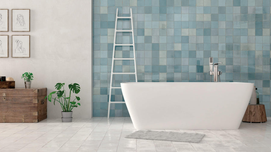 A modern bathroom featuring a white freestanding bathtub, Zellige 5x5 Glossy Subway Tile blue-tiled wall, white ladder, wooden chest, potted plants, framed sketches, and a gray rug on a white tiled floor.