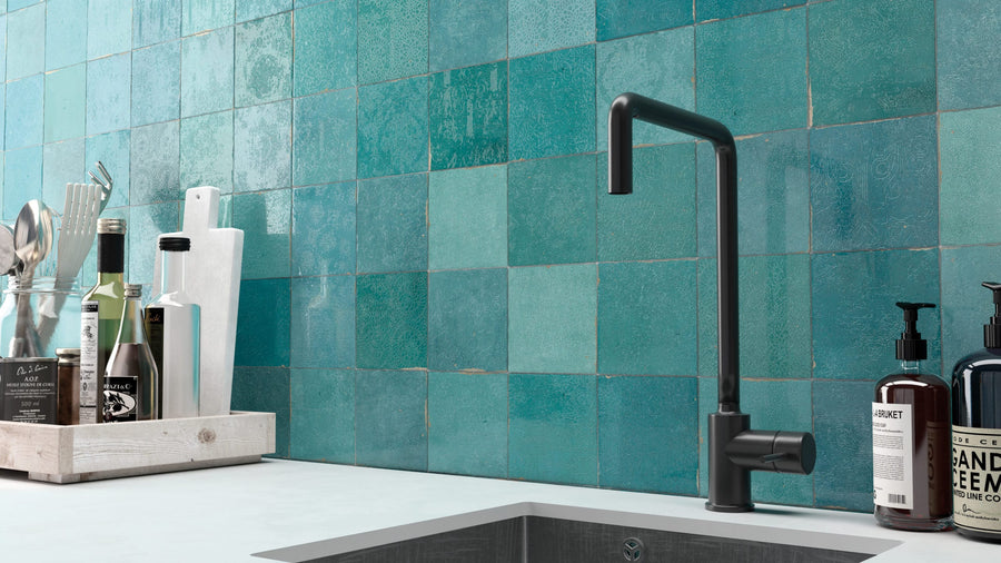 A modern kitchen countertop with a black faucet, stainless steel sink, and bottles of soap and oil. The backsplash features Zellige 5x5 Glossy Subway Tile in turquoise, with varying blue and green shades.