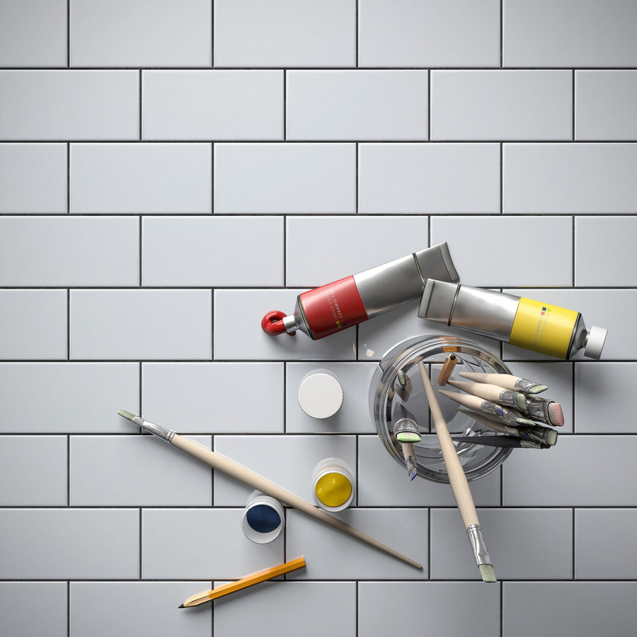 On a U081 3x6 Subway Tile surface, two paint tubes (red and yellow), brushes in a glass jar, two open paint pots (blue and yellow), and a pencil are arranged.