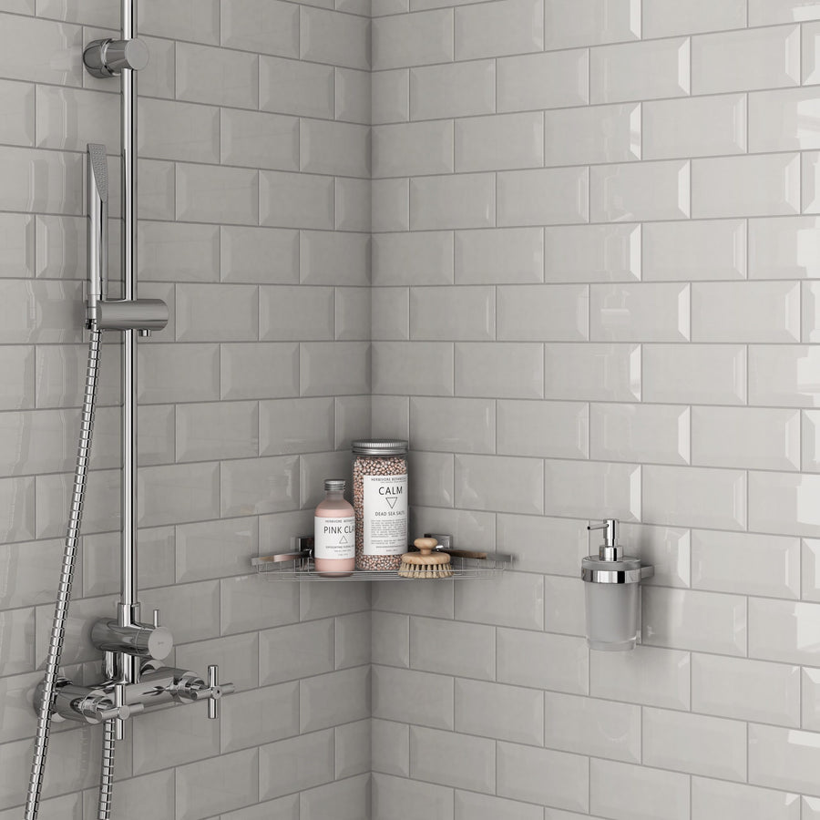 A modern shower corner features U081 3x6 Subway Tile in light gray, a silver showerhead, two glass shelves with bottles and a brush, plus a wall-mounted soap dispenser.