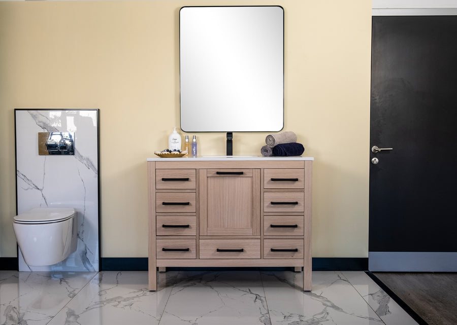 A modern bathroom features a wall-mounted toilet left, the Ashley 42" Vanity in natural oak and a square mirror center, and a dark door right. Elegant white marble tiles cover the floor and lower walls.