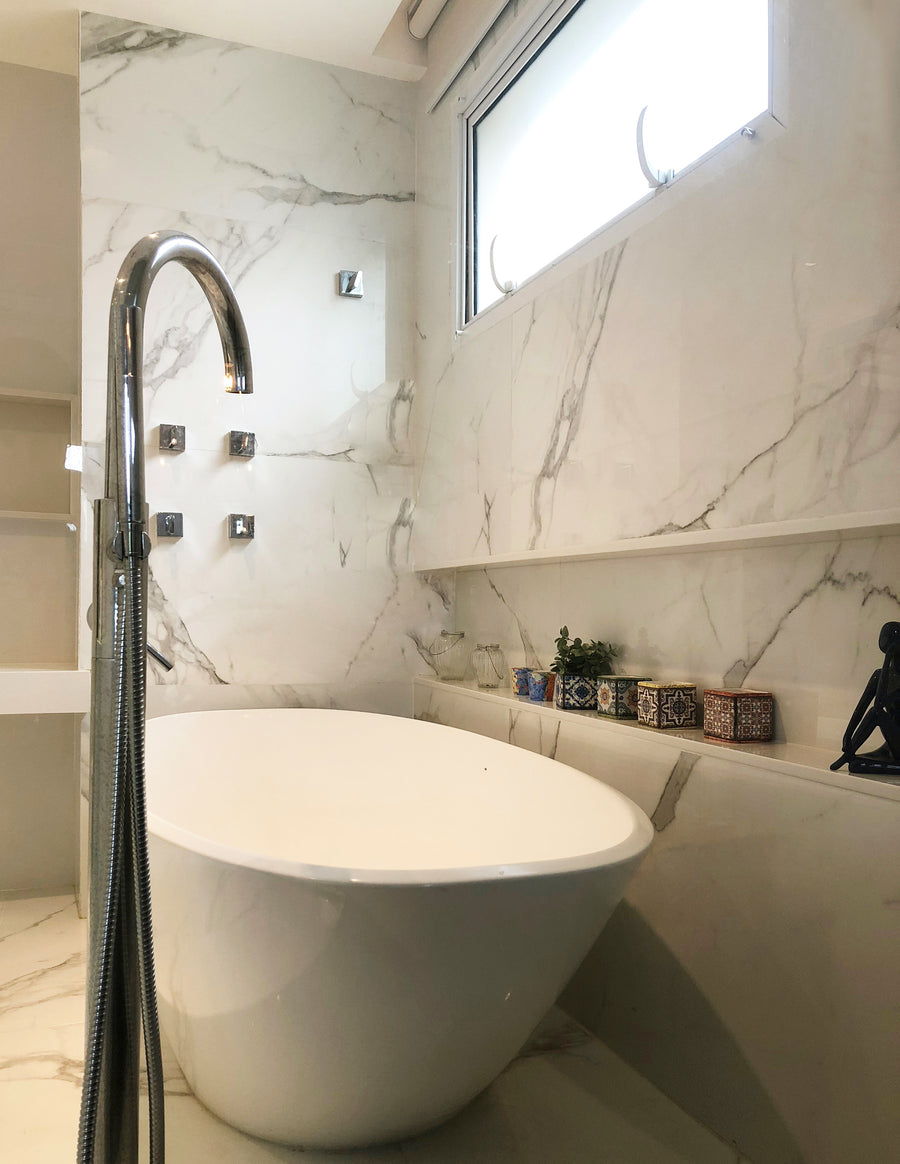 A modern bathroom features Buckingham Palace 24x48 Porcelain Tile on walls and floor, a white freestanding tub with chrome faucet, high window, and shelf with decorative boxes and plant for a serene white and gray design.