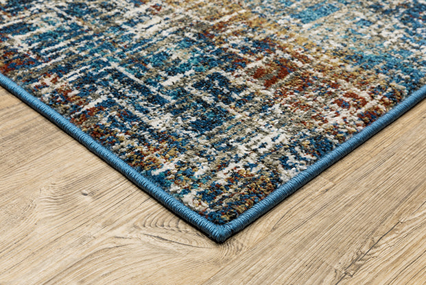 A close-up of the Venice 5573X Area Rug, a multicolored power-loomed polypropylene design with blue, beige, and rust patterns and a neat blue edge, shown on a light wood floor.